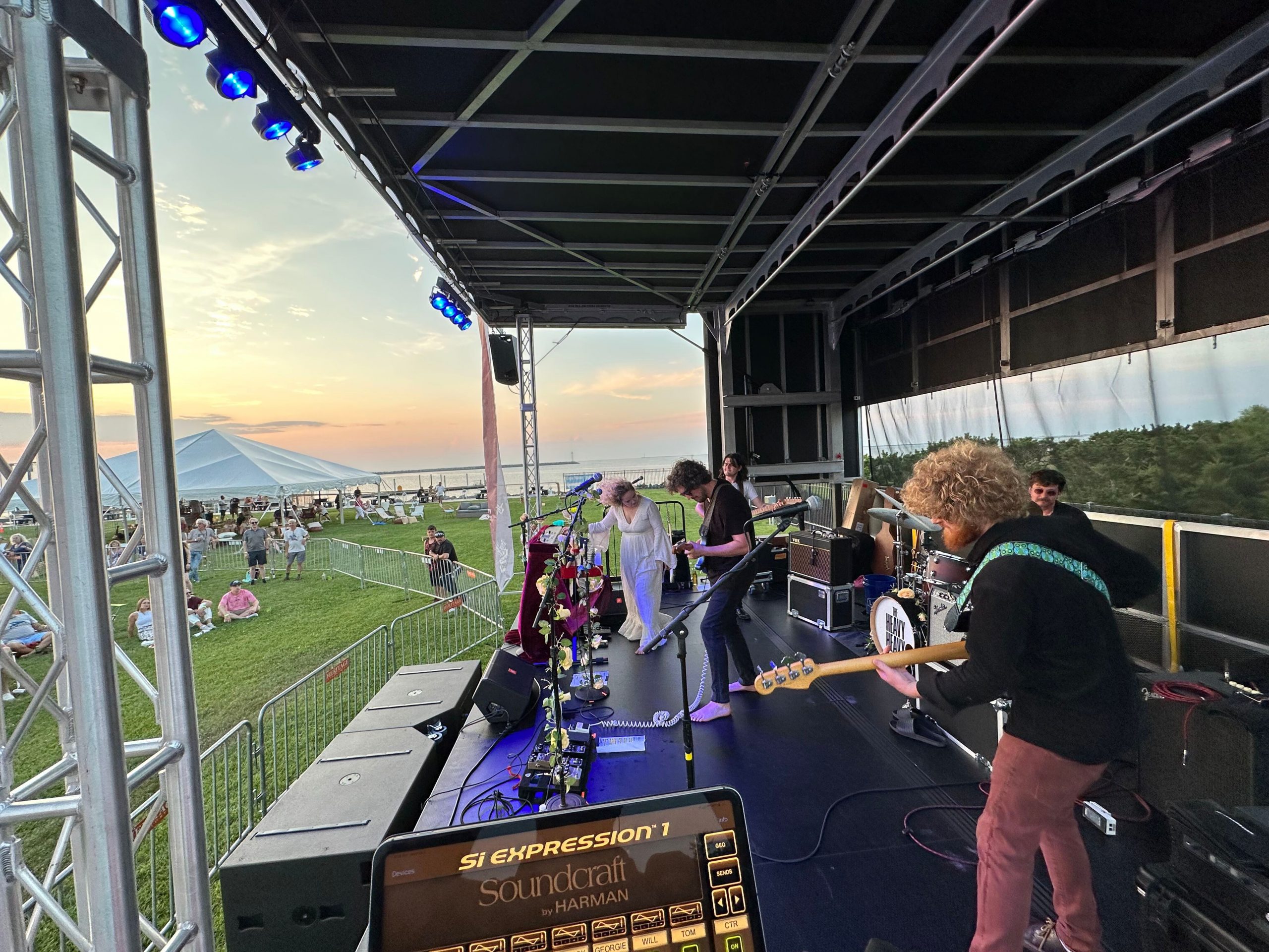 Picture of a band playing the stage from a side perspective. Sunset in the background.